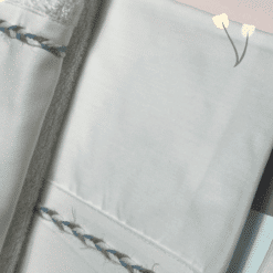 Κοντινή λήψη από ολόλευκο βαπτιστικό σετ με λεπτή πλεκτή κορδέλα σε γαλάζιο και μπεζ πάνω στη φάσα του λαδόπανου και της πετσέτας.