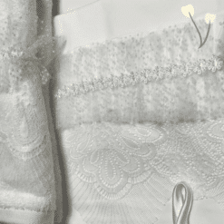 Κοντινή λήψη λευκών βαπτιστικών υφασμάτων με δαντέλα, τούλι με πουά, φιόγκο και λεπτή λουλουδένια τρέσα, σε παστέλ φόντο με δύο γραφικά λουλουδάκια.