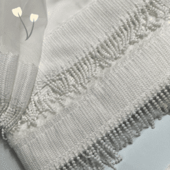 Κοντινή λήψη λευκού υφάσματος και πετσέτας με πυκνή υφαντή τρέσα και κρόσσια, δίπλα σε διάφανο τούλι, πάνω σε παστέλ φόντο με δύο γραφικά λουλουδάκια.