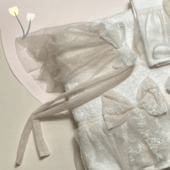 Λεπτομέρειες από εκρού τούλι με λάμψη, λευκή δαντέλα και φιόγκους πάνω σε λευκή πετσέτα, με παστέλ φόντο και μικρά γραφικά λουλουδάκια.