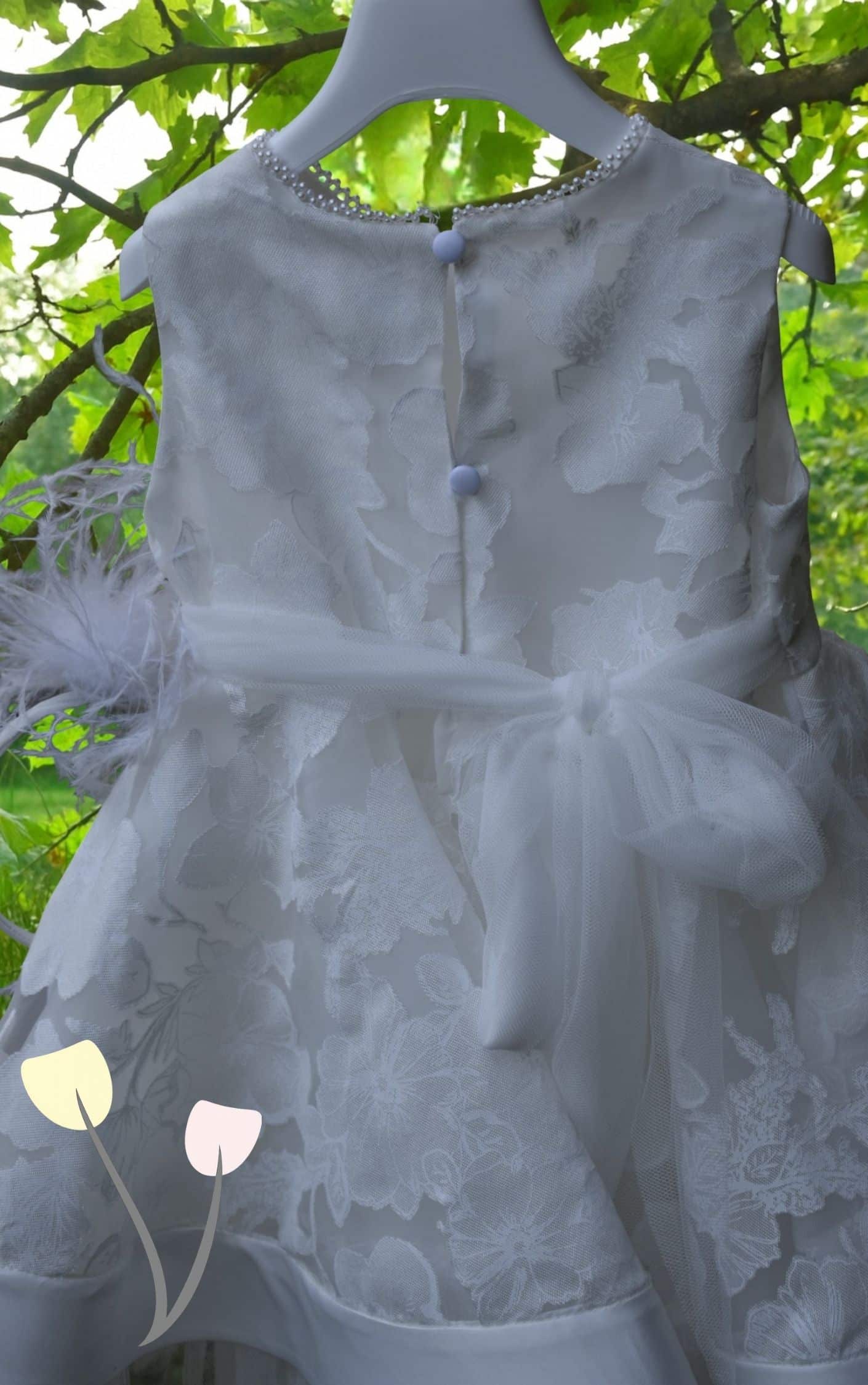 Πίσω όψη ιβουάρ βαπτιστικού φορέματος με ανάγλυφα λουλούδια, σειρά από πέρλες στο ντεκολτέ, δύο κουμπάκια στην πλάτη και ζώνη από τούλι δεμένη σε φιόγκο, κρεμασμένο σε κλαδί μέσα στη φύση.