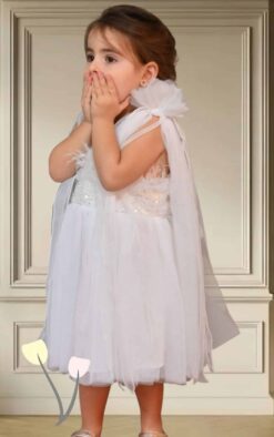 Βαπτιστικό ρούχο κορίτσι Θεσσαλονίκη Baby Lovezu Μικρό κορίτσι με λευκό φόρεμα από τούλι και ζώνη με παγιέτες, κρατά τα χέρια στο στόμα σαν έκπληξη, σε ανοιχτόχρωμο κλασικό φόντο