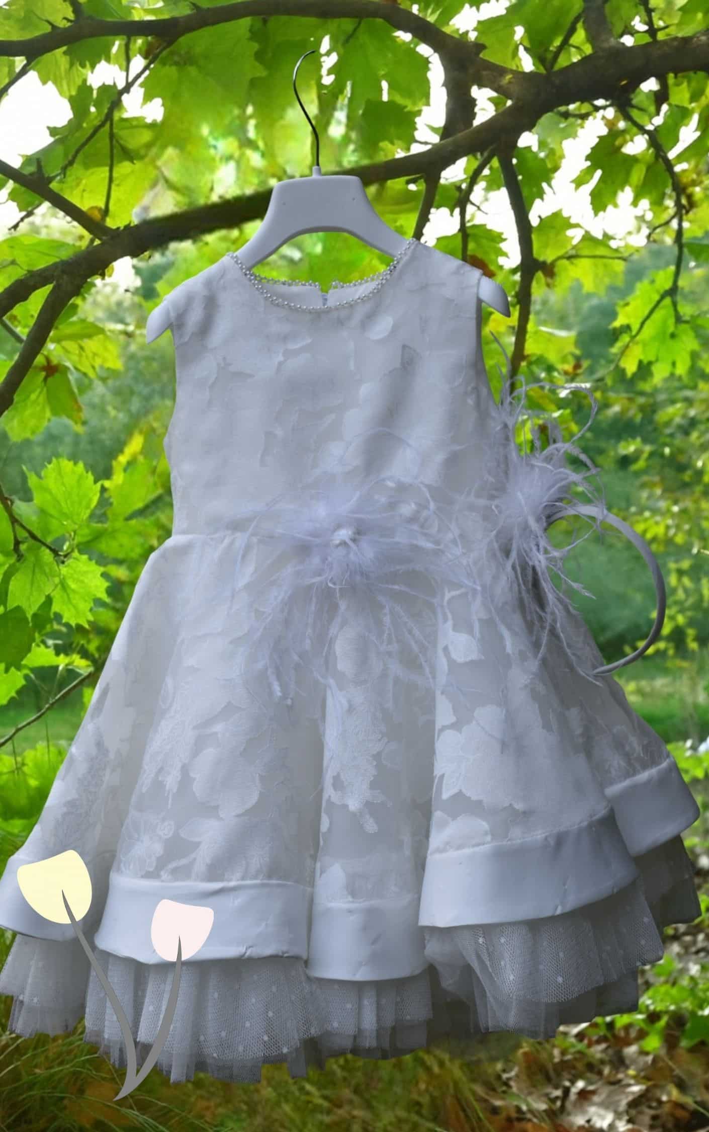 Ιβουαρ βαπτιστικό φόρεμα κοριτσιού με ανάγλυφα λουλούδια, πέρλες στο ντεκολτέ, φτερά στη μέση και διπλή φούστα με τούλι, κρεμασμένο σε κλαδί μέσα στη φύση.