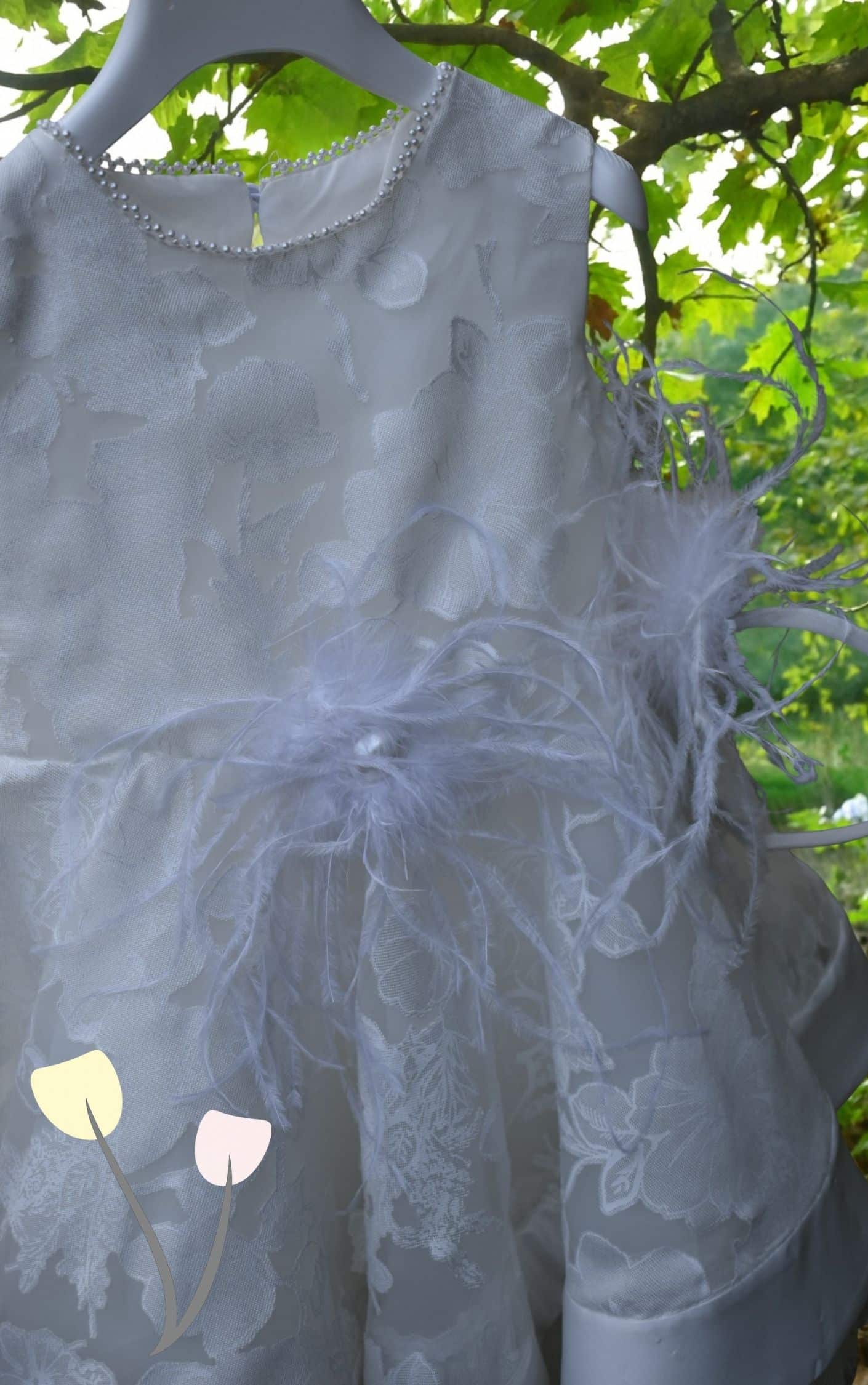 Κοντινό σε ιβουάρ βαπτιστικό φόρεμα με ανάγλυφα λουλούδια, πέρλες στο ντεκολτέ και διακοσμητικό από απαλό φτερό στη μέση, κρεμασμένο σε κλαδί μέσα στη φύση.